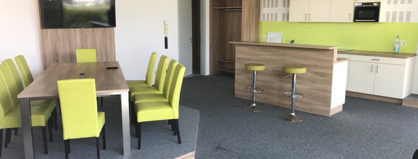 Besprechungsraum im Co-Working+ mit Sesseln, Tisch und Flatscreen im linken Bildteil, Küchenzeile mit Theke im rechten Bildteil.